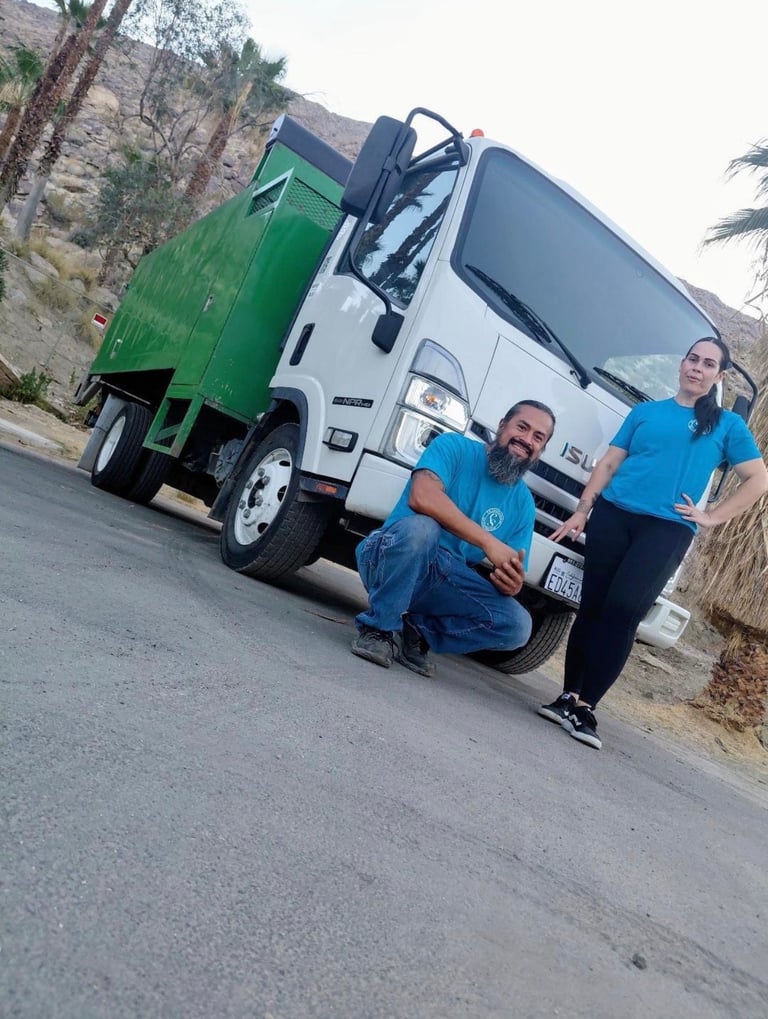 Three workers in blue uniforms standing in front of a white garbage collection truck with a green waste bin attached