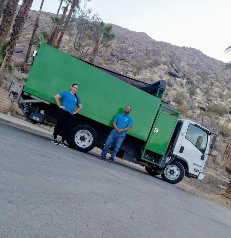 Two workers in blue shirts standing beside a green and white garbage truck on a residential street with rocky hillside in background